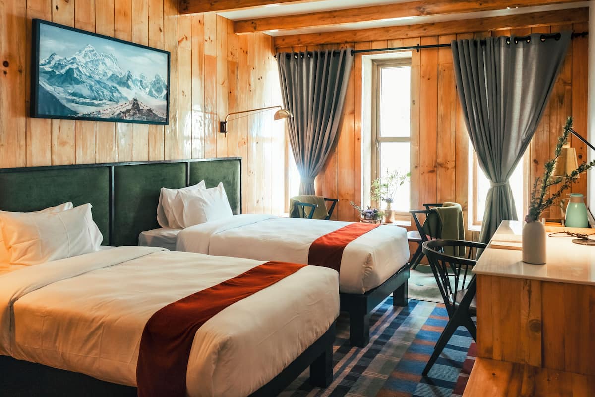 Habitación de hotel con dos camas, un escritorio, una silla y un ventanal con cortinas.