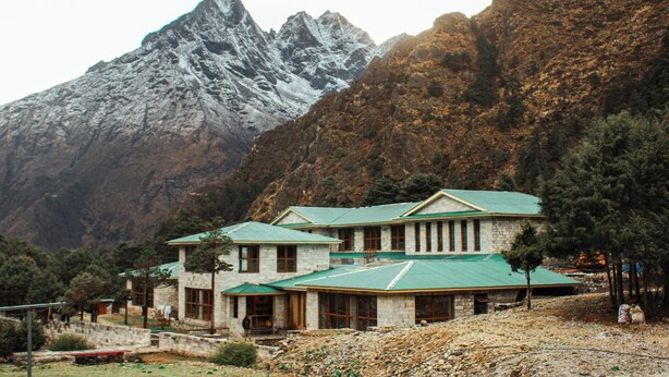 A stone building with a green roof, surrounded by trees and mountains.