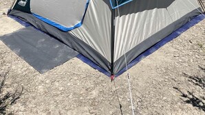 A dome-shaped tent with a gray and blue color scheme, set on a rocky ground with sparse vegetation in the background.