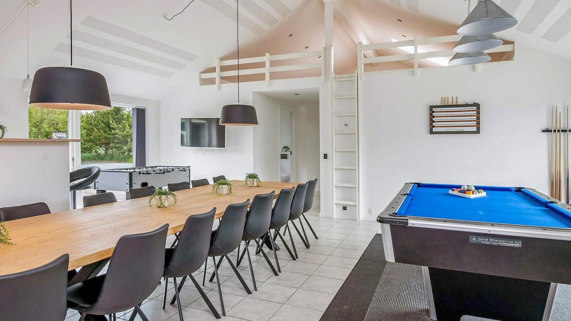 A modern room with a pool table, a dining area with a long table and chairs, a foosball table, and a television mounted on the wall.