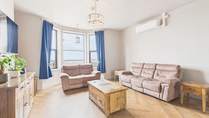 Una sala de estar con un sofá color beige, una mesa de centro de madera y una mesita auxiliar de madera. Se ve una ventana con cortinas azules, una planta en maceta y una lámpara colgante.