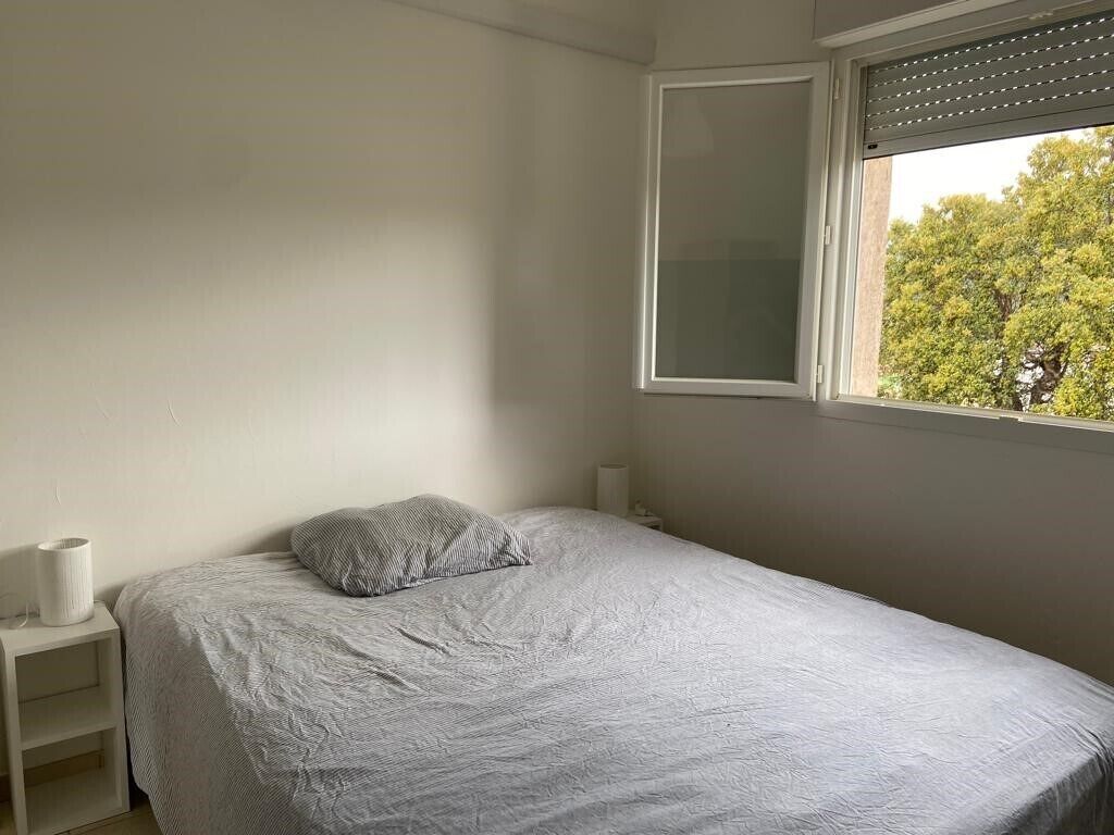 Una cama individual con colcha gris y una almohada, una mesita de noche con lámpara, y una ventana con persianas.