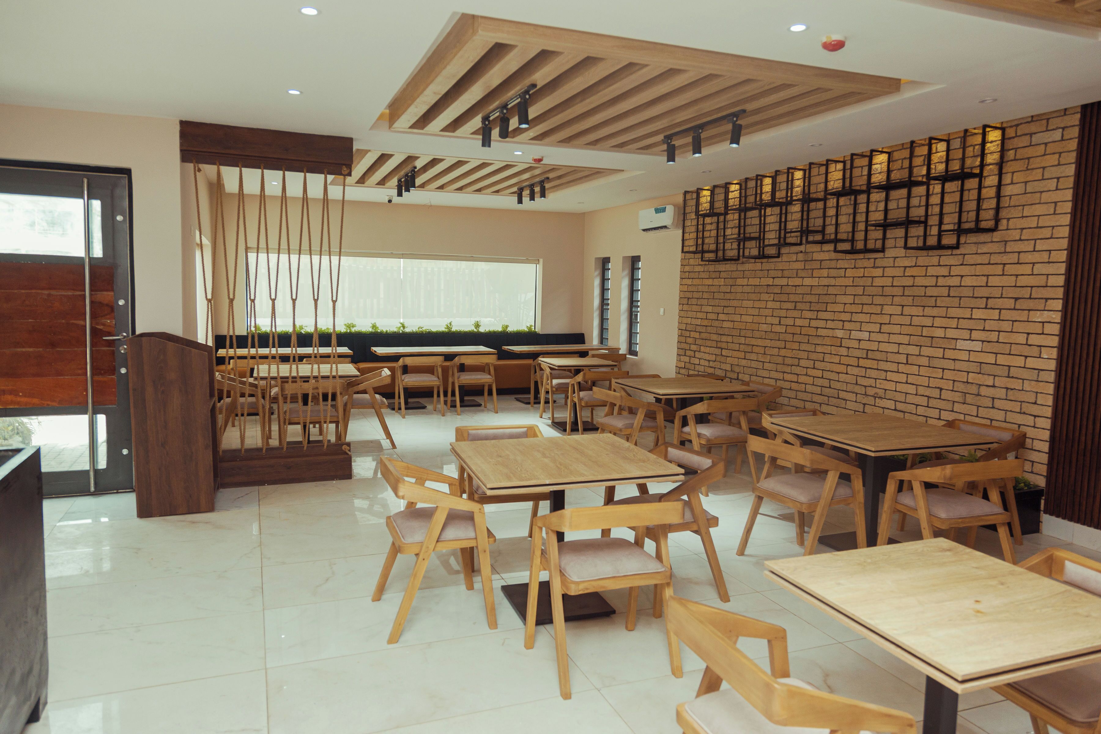 A spacious restaurant with wooden tables and chairs, a brick wall, and a modern ceiling design.