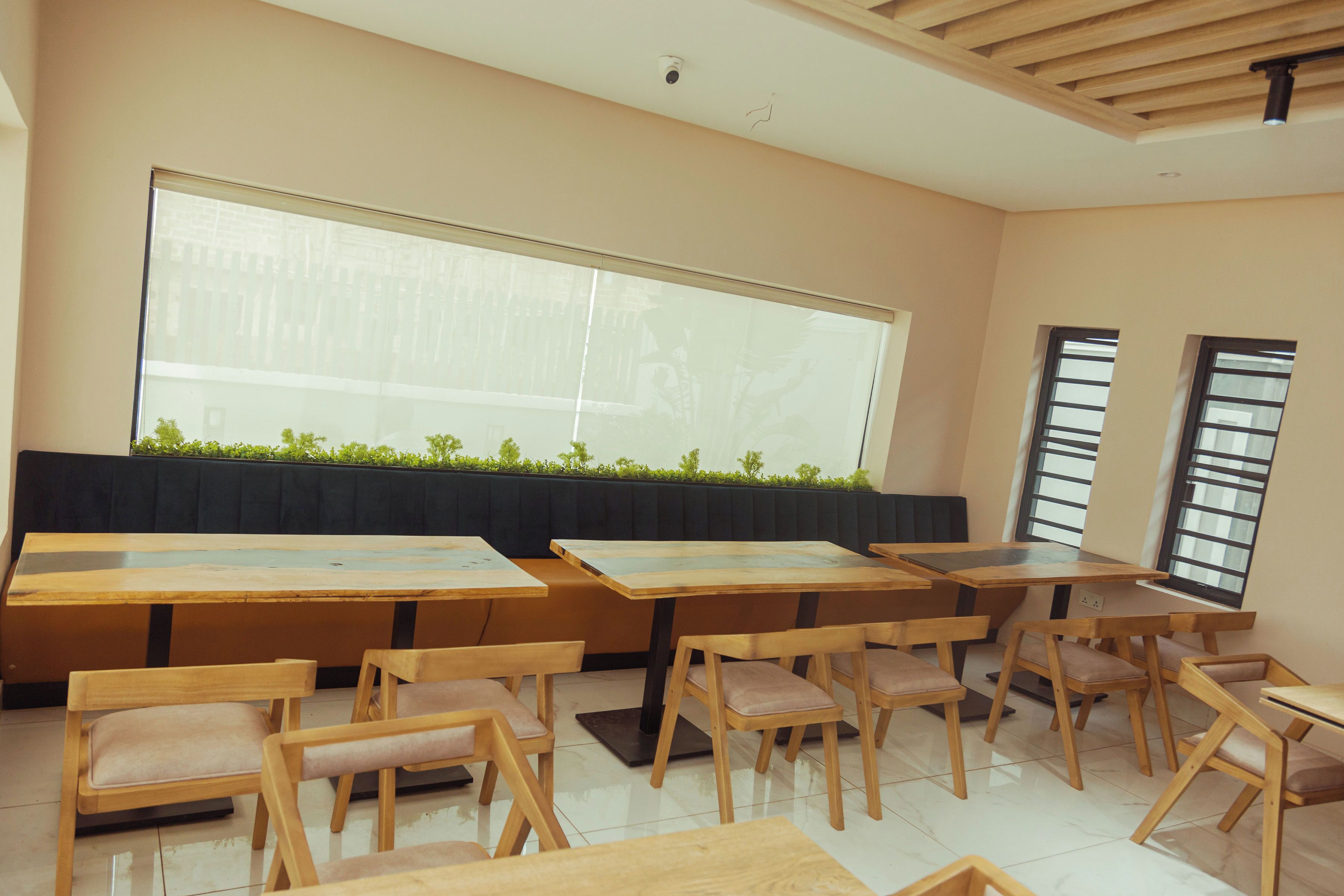 A modern dining area with wooden tables and chairs, large windows with blinds, and a green plant wall.