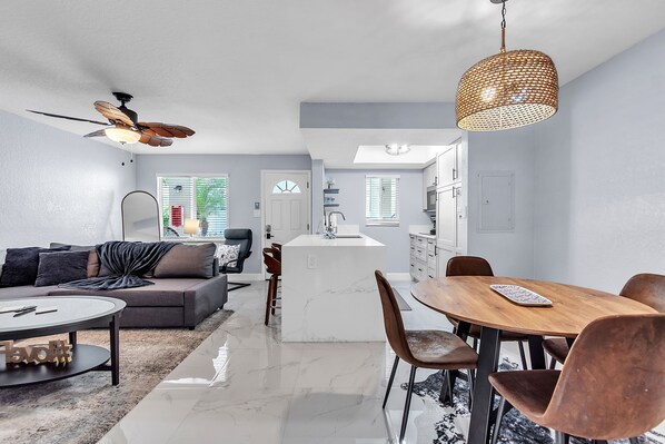 A modern kitchen with a central island, a dining area with a wooden table and chairs, a living room with a sofa and coffee table, and a ceiling fan.