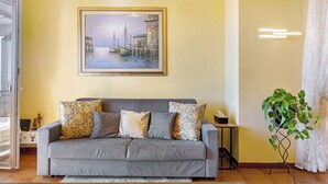 Ein Wohnzimmer mit grauem Sofa, einem gerahmten Gemälde und einem Blumentopf.