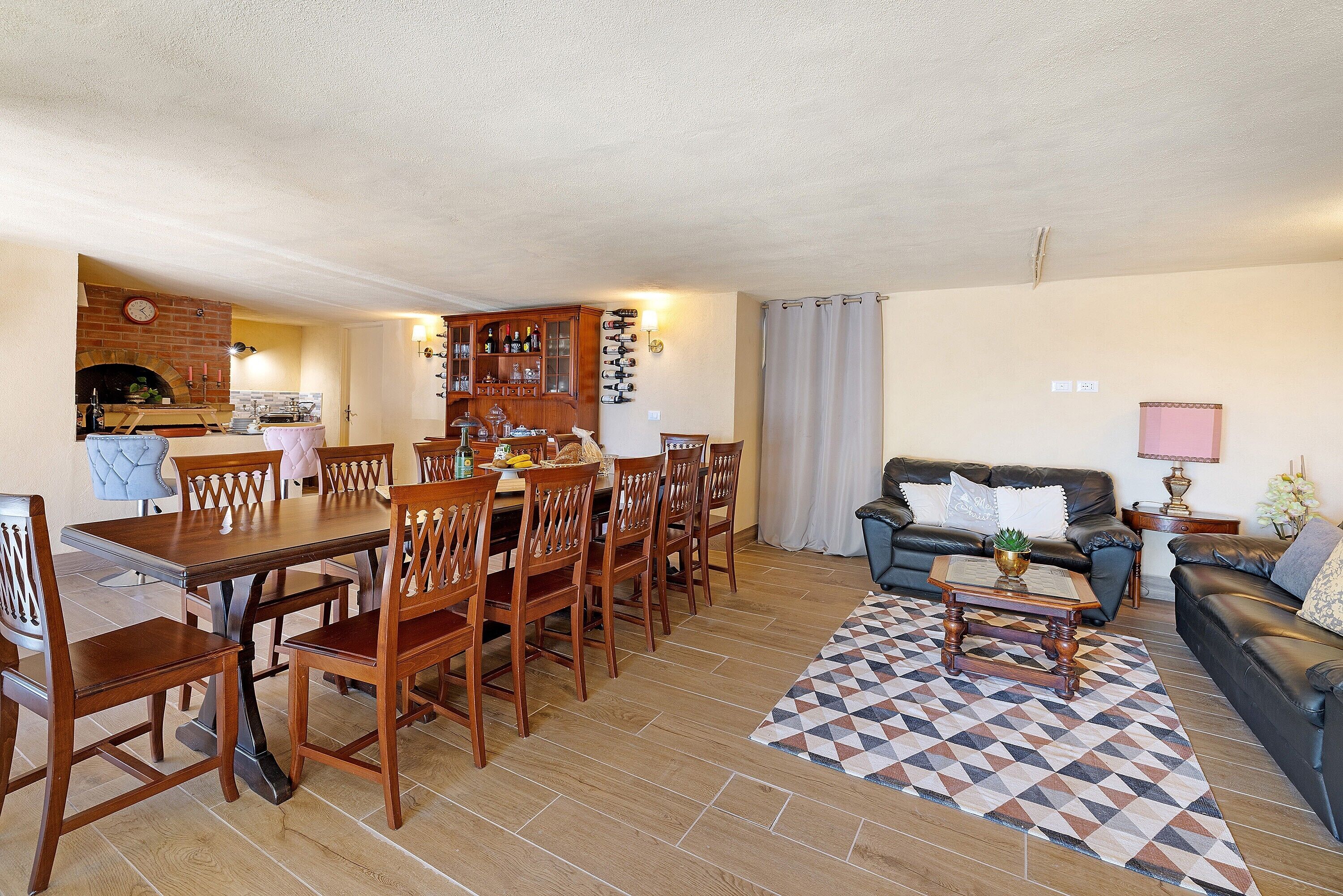 A dining area with a wooden table and chairs, a kitchen with a brick wall and a clock, a living room with a sofa and a patterned rug.