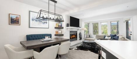 A modern living room with a fireplace, a dining area with a wooden table and white chairs, a blue sofa, and a large window with a view of the city.