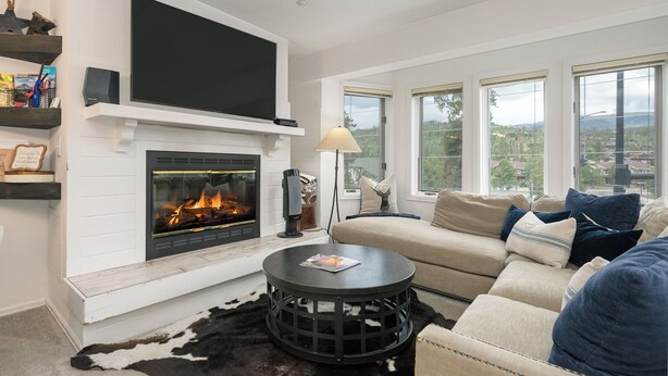 A living room with a fireplace, a television mounted above it, a sofa with cushions, and large windows offering a view of the outdoors.