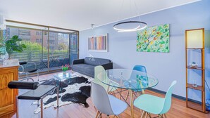 A modern living room with a glass coffee table, a sofa, a dining area with chairs, a bookshelf, and artwork on the walls. - Urban Bloom 3D, Wifi y Estacionamiento (Ñuñoa)
