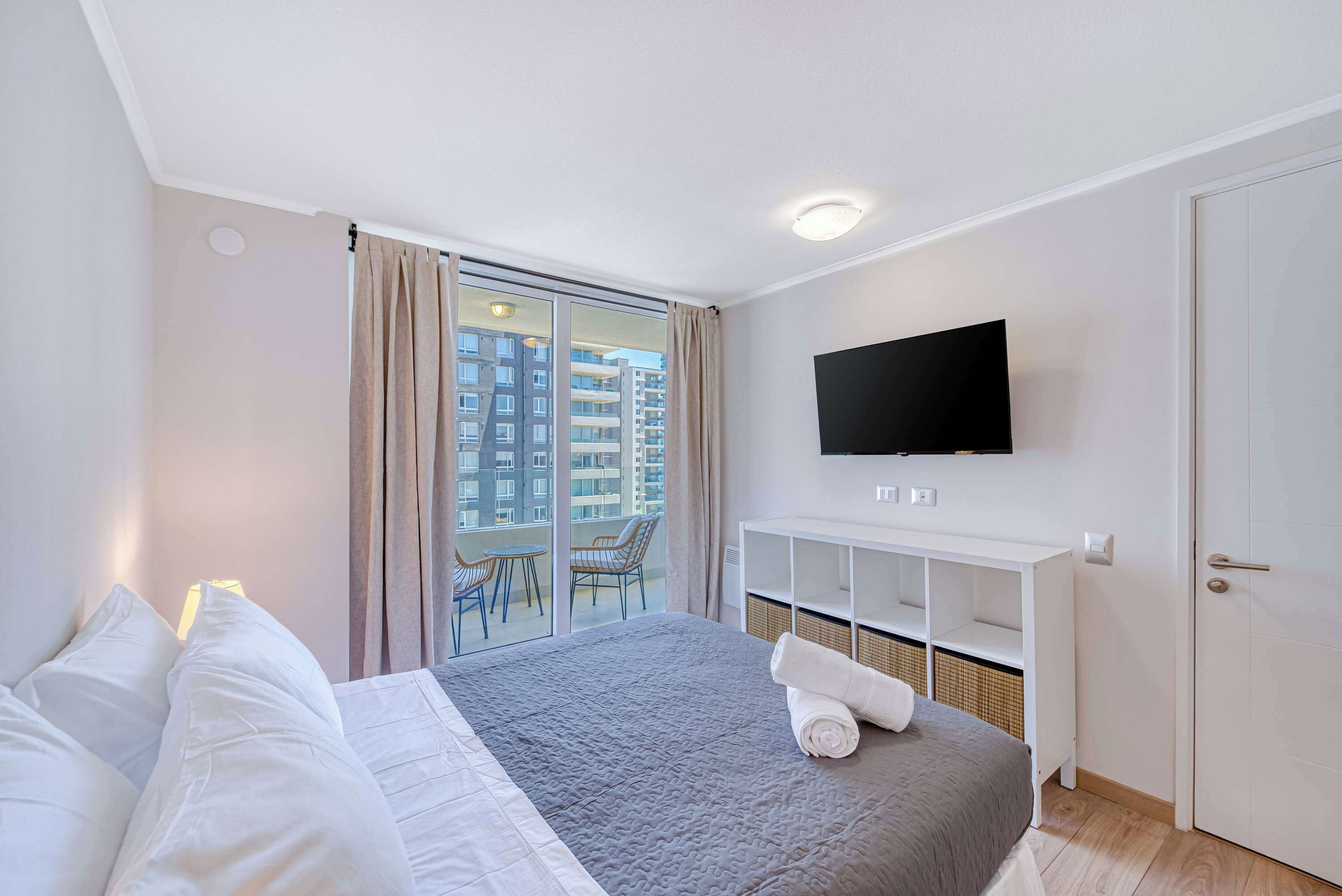A hotel room with a bed, a TV, a balcony with chairs, and a white wardrobe.