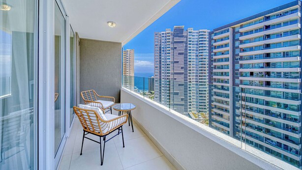 A balcony with wicker chairs and a small table, overlooking high-rise buildings and a clear sky. - Vista Infinita (Concón)