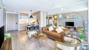 Uma sala de estar moderna com sofá, mesa de centro e cadeiras, cozinha ao fundo e televisão de tela plana.