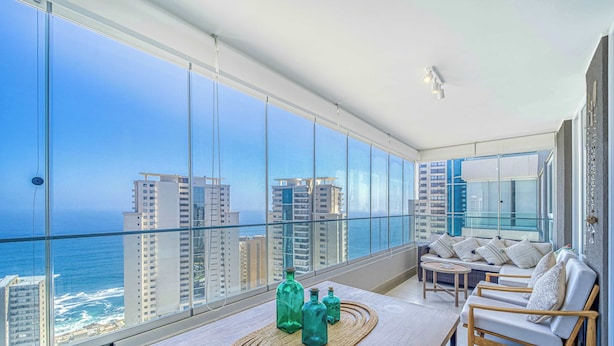 Varanda com vista para o oceano e para a cidade, com uma mesa de madeira, garrafas decorativas, um sofá e uma pequena mesa redonda.