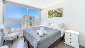 Quarto de hotel com uma janela grande, uma cama com colcha cinza estampada, uma cadeira branca, uma mesinha de cabeceira com abajur e um quadro na parede.