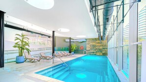 Piscina interna com espreguiçadeiras, uma planta em vaso e vista para um edifício.