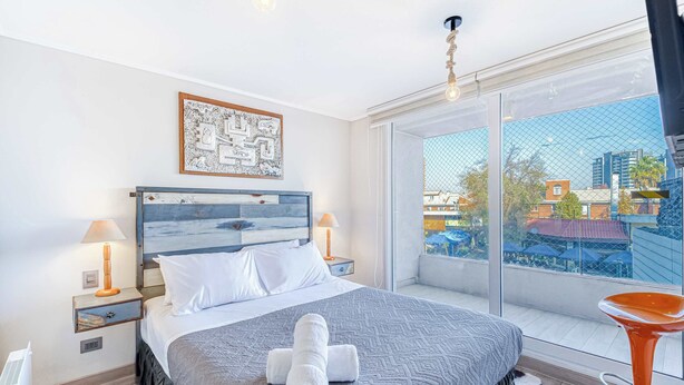 Una habitación de hotel con cama, mesita de noche, lámpara, ventana con vista a edificios y una silla.