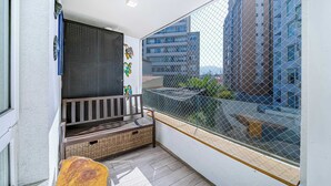 Un balcón con un banco de mimbre, una parrilla de metal y vistas de edificios a través de una malla.
