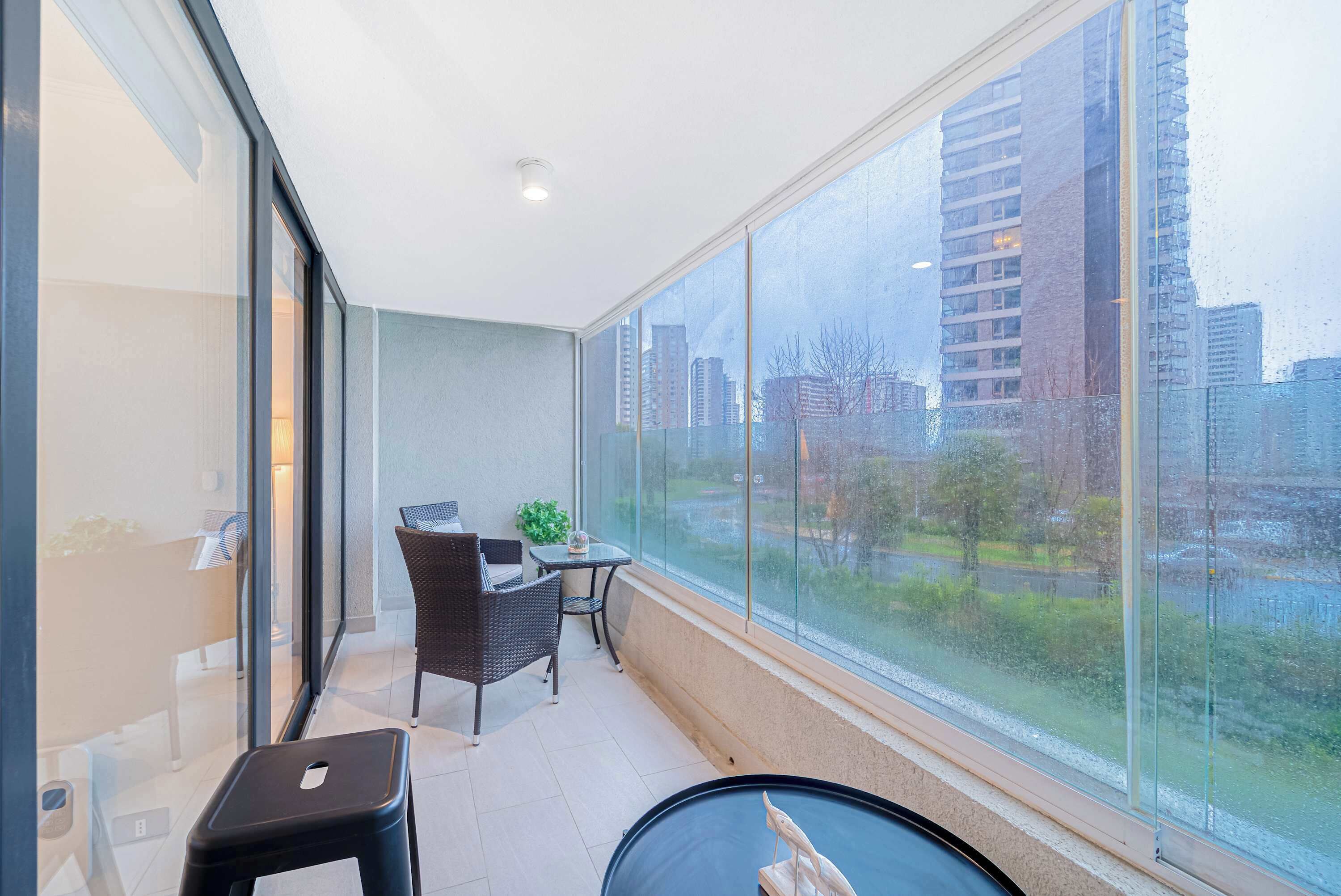 A balcony with a table and chairs, a small stool, and a view of a cityscape through large windows.