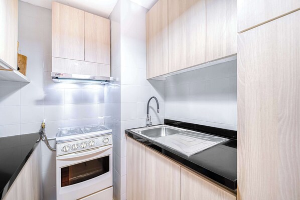 A compact kitchen with a stove, oven, and sink, light wood cabinets, and white tiled walls.