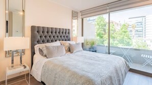 Une chambre moderne avec une tête de lit capitonnée, un grand miroir et un balcon offrant une vue sur les immeubles et les arbres.