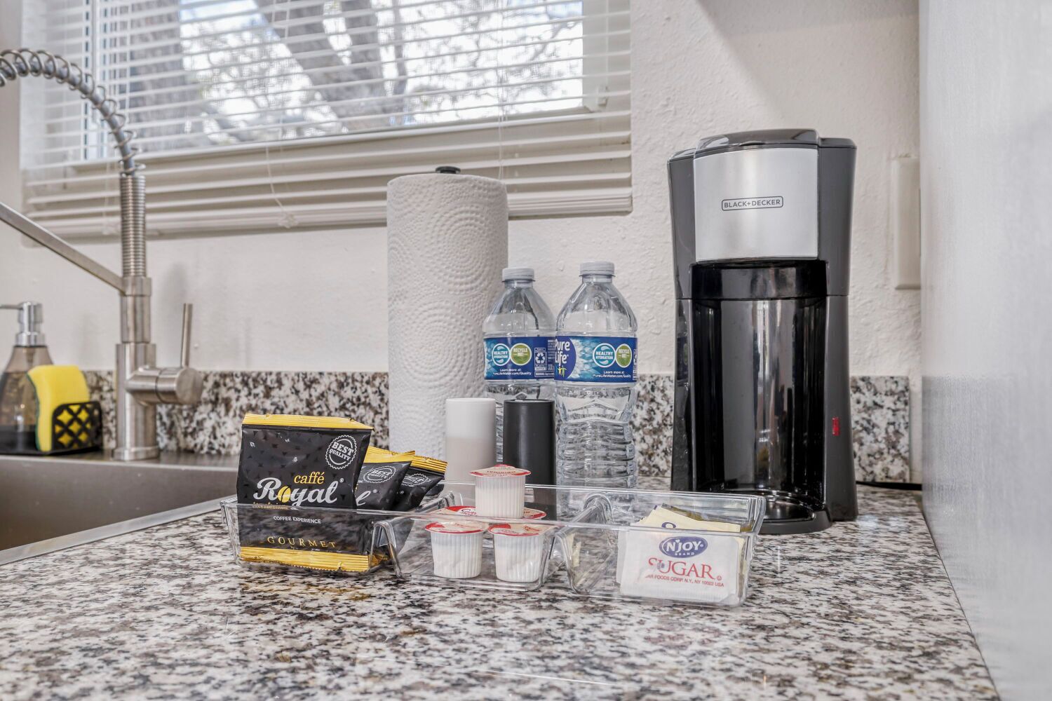 Eine Küchenarbeitsplatte mit Kaffeemaschine, Flaschenwasser, Kaffeekapseln, Zuckerportionen und einem Papiertuchspender.