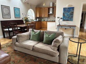 Ein gemütliches Wohnzimmer mit einem beigefarbenen Sofa, einem Holzesstisch und einer Küche im Hintergrund.