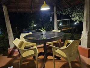 A round table with four wicker chairs, a white mug, and a yellow pendant light in an outdoor setting at night. - Turmerica Resorts (Kalpetta)