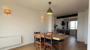 A dining area with a wooden table and chairs, a kitchen in the background, and a ceiling light fixture. - Beachfront Apartment w/ Balcony & Sea Views (Valras-Plage)
