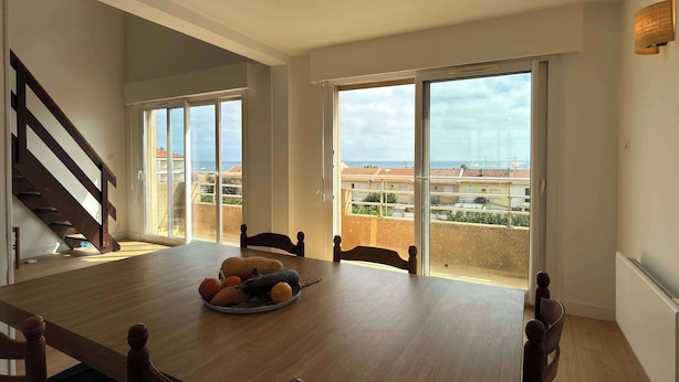 A dining area with a wooden table, chairs, and a fruit bowl. There is a staircase on the left and a balcony with a view of buildings and the sea. - Beachfront Apartment w/ Balcony & Sea Views (Valras-Plage)