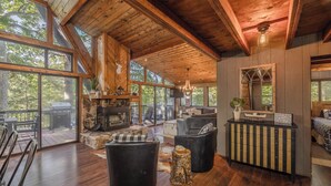 A spacious living room with a stone fireplace, wooden ceiling beams, and large windows offering a view of the outdoors.