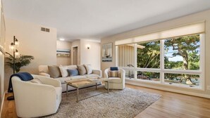 Ein modernes Wohnzimmer mit einer Couch, Sesseln, einem Couchtisch und einem großen Fenster mit Blick auf Bäume und einen Balkon.