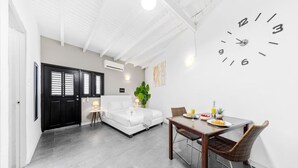 Een moderne hotelkamer met een wit bed, een eettafel met stoelen, een grote wandklok en een deur met een zwarte lijst.