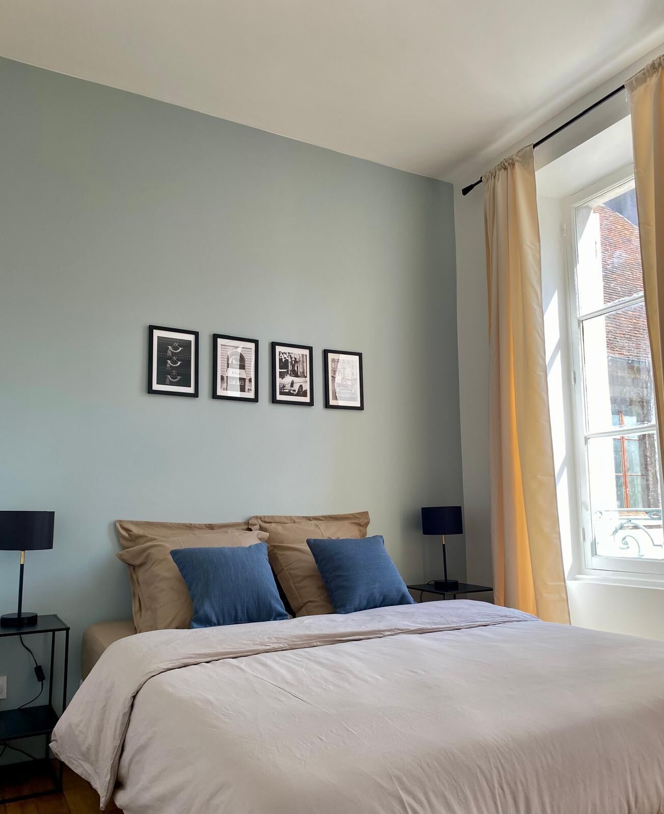 Une chambre à coucher avec un lit, deux tables de chevet, une fenêtre avec des rideaux et trois tableaux encadrés accrochés au mur.