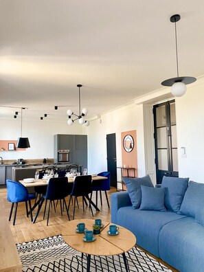 A modern living room with a blue sofa, a dining area with a table set for four, and a kitchen in the background.