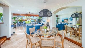 A spacious living area with a glass-top dining table surrounded by wicker chairs, a ceiling fan, and a large window offering a view of the outdoors.