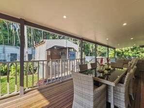 Une terrasse extérieure couverte, aménagée avec des meubles en osier, un espace barbecue et une vue sur une maison et des arbres.