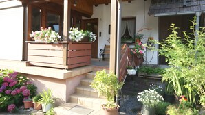 A wooden balcony with potted plants and flowers, a small outdoor seating area, and a staircase leading to a door.