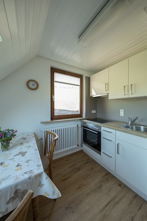 A compact kitchen with a window, a radiator, a table with a floral tablecloth, and wooden chairs.