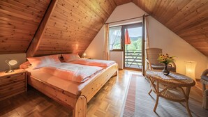 A bedroom with a wooden ceiling, a bed, a nightstand, a table with a vase of flowers, and a view of greenery outside. - Studio 'Kornblümchen' with private terrace, shared garden and Wi-Fi (Oberharmersbach)