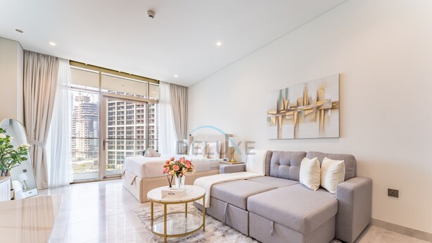 A spacious living room with a grey sofa, a round coffee table, and a large window with a view of buildings. - Furnished Studio with Burj Khalifa View, Near Dubai Water Canal (Dubai)