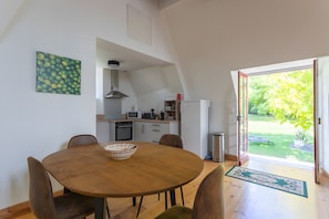 Un coin repas avec une table ronde en bois, des chaises, un saladier, et une vue sur la cuisine et le jardin, grâce à une porte ouverte.