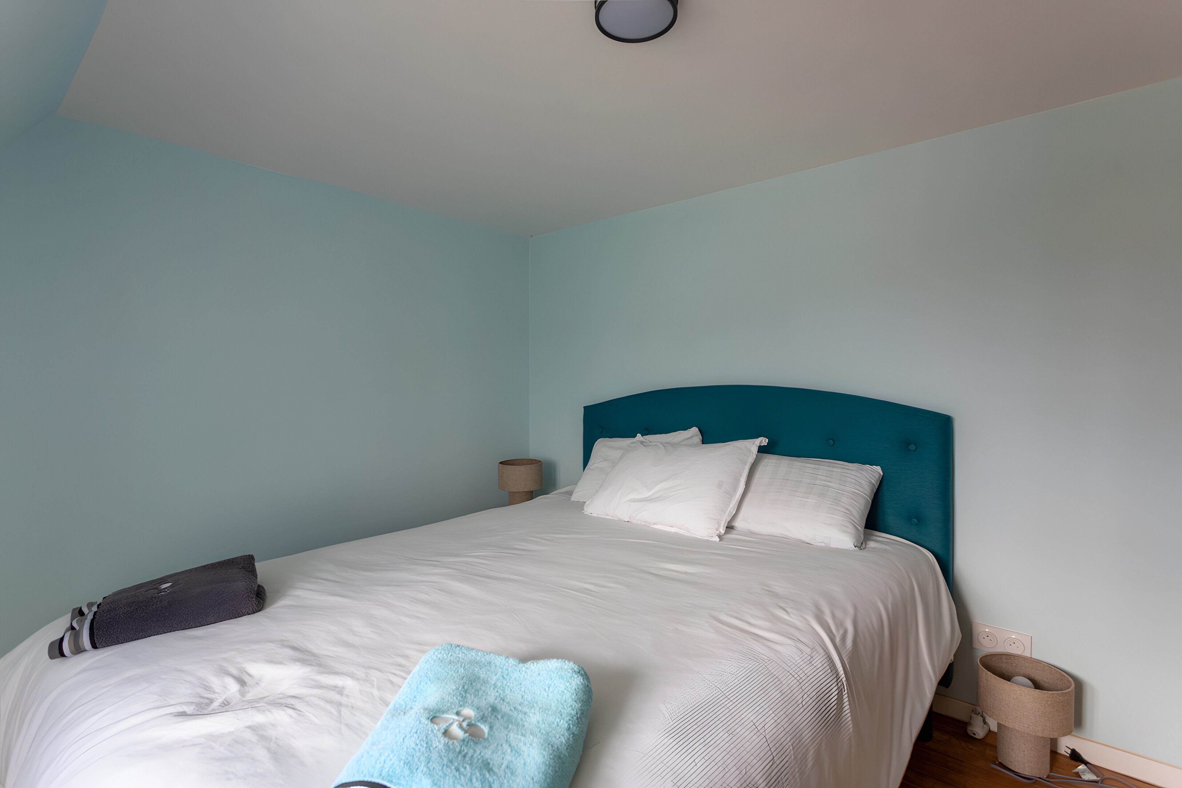A neatly made bed with a teal headboard, a white pillow, a blue throw pillow, and a dark-colored bag on the left side.
