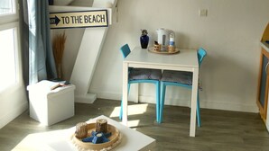 A room with a staircase, a table with chairs, a surfboard decoration, and a sign reading 'TO THE BEACH'.