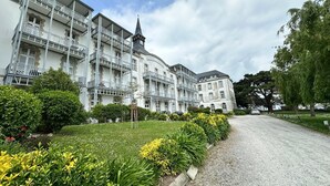 A historic building with multiple balconies and a tower, surrounded by well-maintained gardens and a gravel driveway.