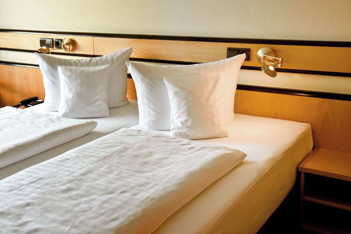 Une chambre d’hôtel avec deux lits, chacun étant équipé de draps et d’oreillers blancs. Il y a une tête de lit en bois avec un luminaire et une table de chevet avec une lampe.