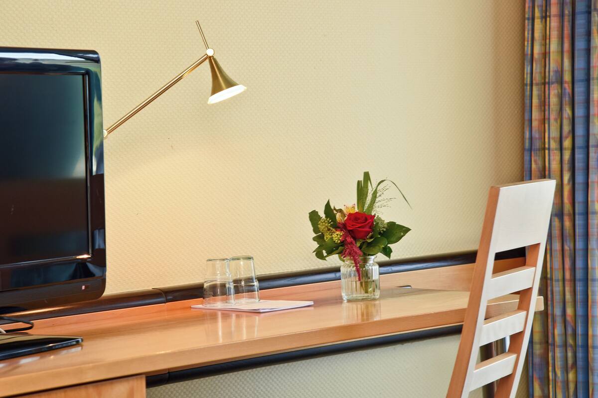 Une chambre d’hôtel avec un bureau en bois, une télévision, une lampe de bureau, un vase avec des fleurs et une chaise.