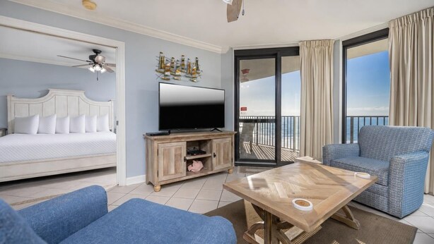 A hotel room with a bed, a TV, a wooden coffee table, a blue sofa, and a balcony with a view. - Phoenix Vi 61004 2 Bedroom Condo (Orange Beach)