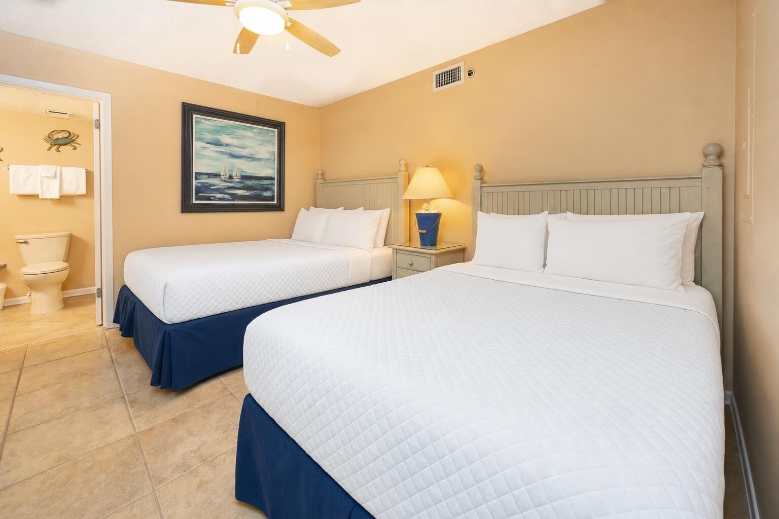 A hotel room with two beds, a ceiling fan, a painting, a nightstand with a lamp, and a view of the sea.