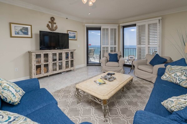 A living room with a television, two sofas, a coffee table, and a view of the ocean through large windows. - Phoenix Vi 61103 3 Bedroom Condo (Orange Beach)